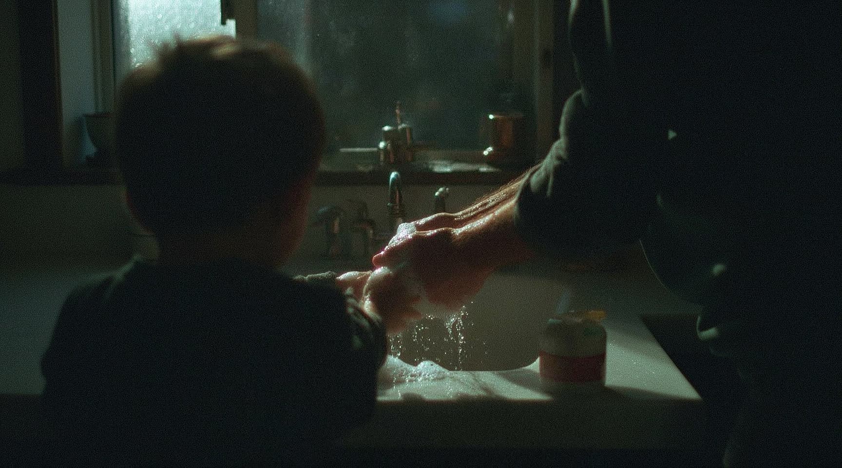 A parent washing a child’s hands at a kitchen sink by the window, soap catching the evening light